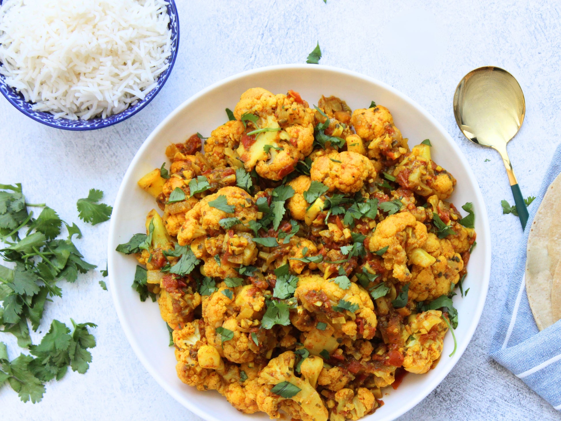 Cauliflower florets cooking with golden turmeric and chopped red tomatoes and sprinkled with fresh green cilantro. Blue bowl of long grain Basmati rice. Chaptis wrapped in a blue and white striped kitchen towel Gold spoon with green handle tucked behind towel.